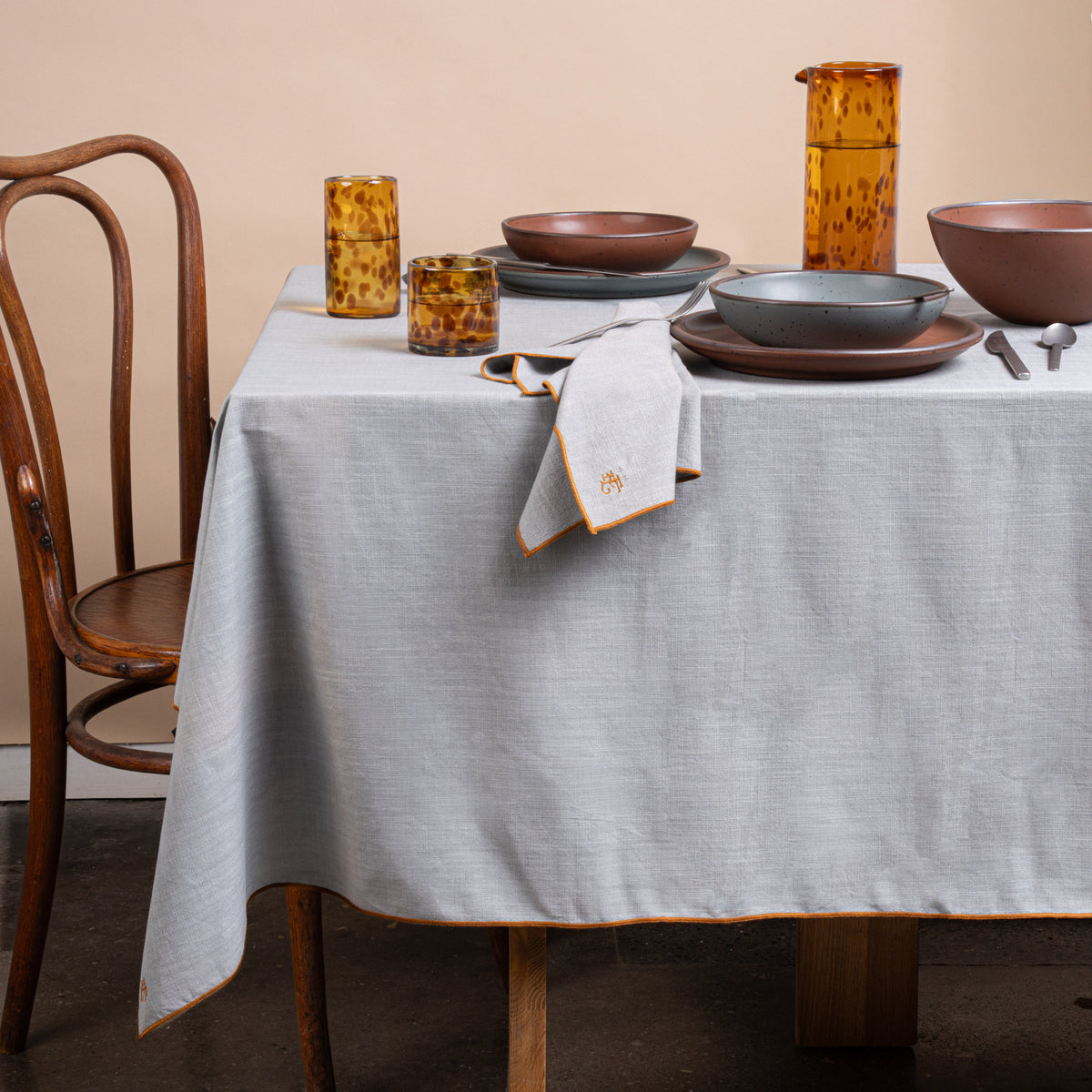 A table set with ceramic pottery, glasses of water, silverware, and a light blue table linen with rust-colored piping and embroidered monogram.