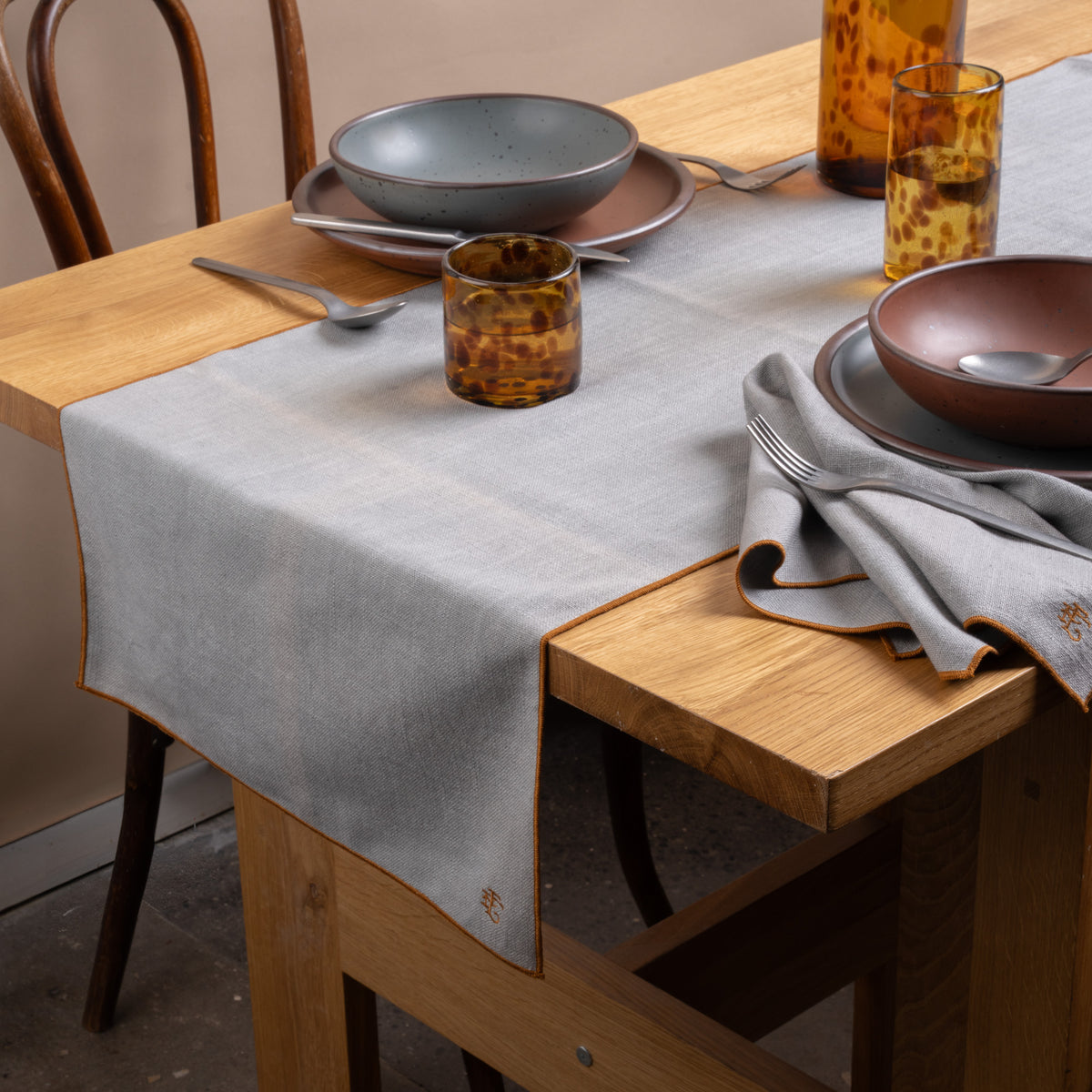 A table set with ceramic pottery, glasses of water, silverware, and a light blue table linen with rust-colored piping and embroidered monogram.