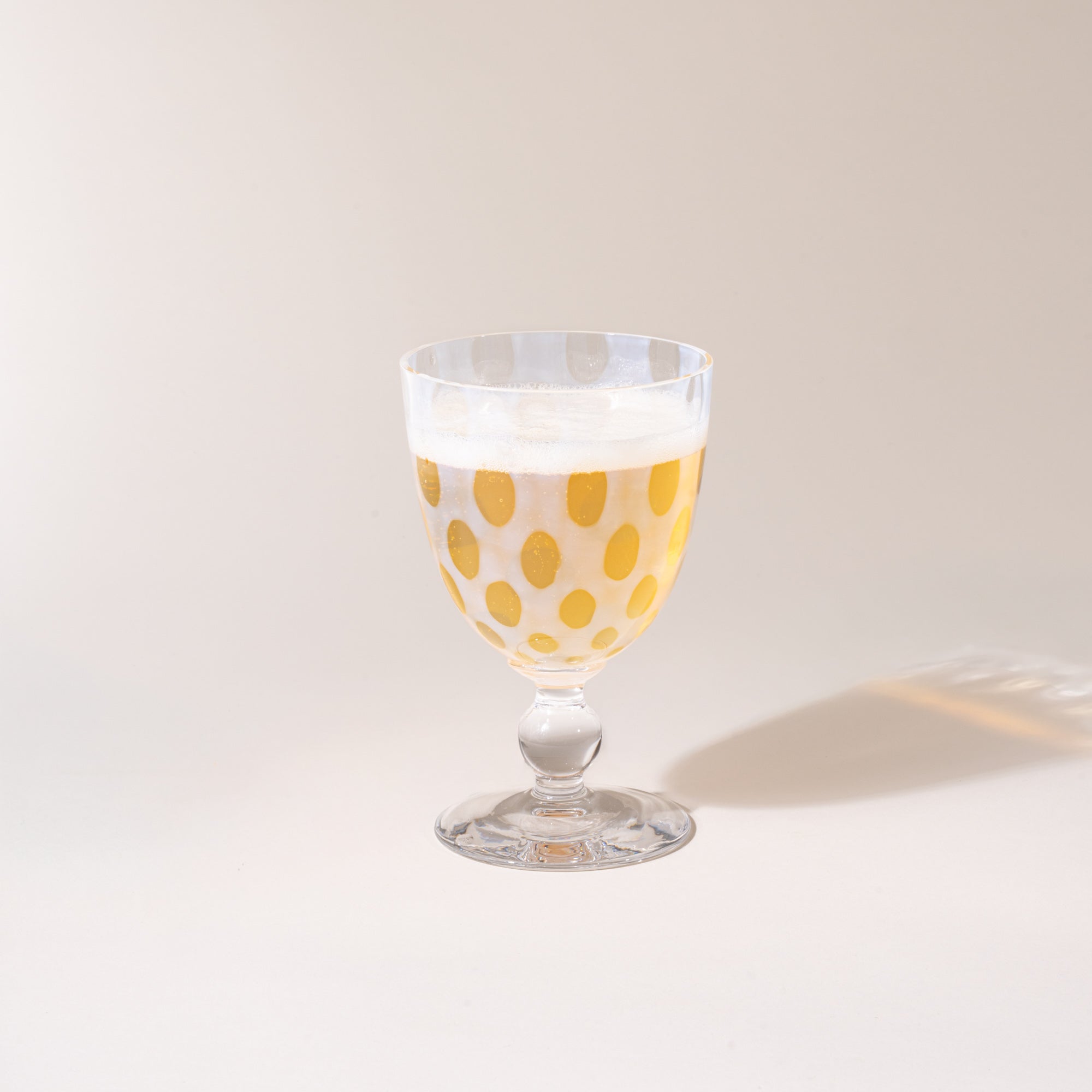 A clear stemmed goblet with a subtle polka-dot pattern, photographed against a warm neutral background with a soft cast shadow, with a sparkling drink inside.