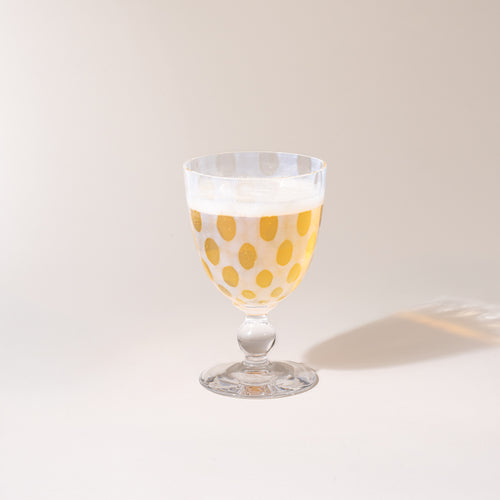 A clear stemmed goblet with a subtle polka-dot pattern, photographed against a warm neutral background with a soft cast shadow, with a sparkling drink inside.
