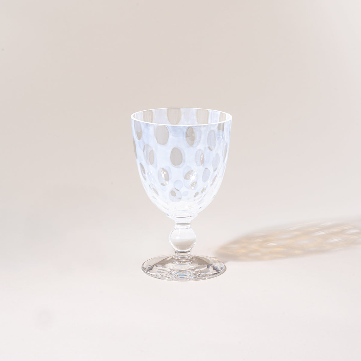 A clear stemmed goblet with a subtle polka-dot pattern, photographed empty against a warm neutral background with a soft cast shadow.