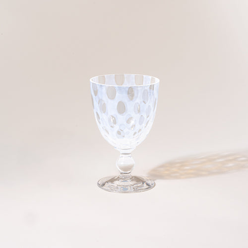 A clear stemmed goblet with a subtle polka-dot pattern, photographed empty against a warm neutral background with a soft cast shadow.