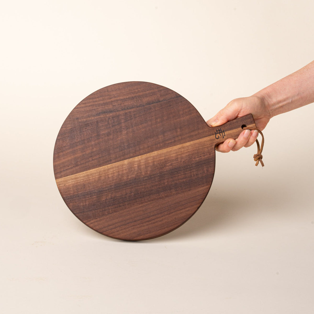 A hand holds a small round walnut serving board with a short handle and leather hanging loop, shown held against a warm cream background.
