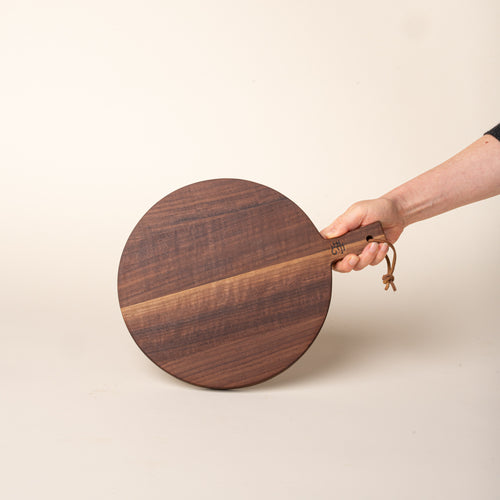 A hand holds a small round walnut serving board with a short handle and leather hanging loop, shown held against a warm cream background.