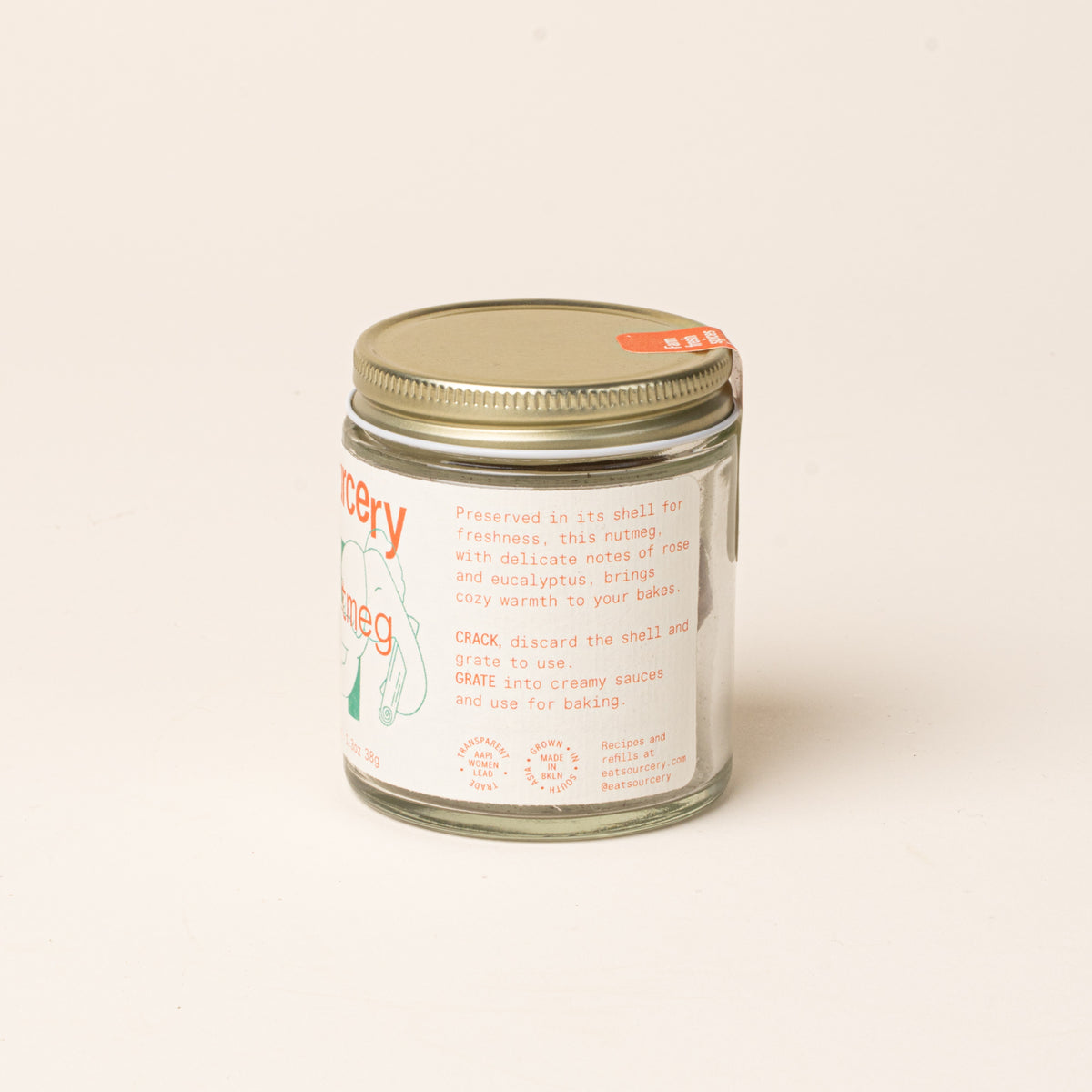Side of a glass jar of Sourcery nutmeg with a gold metal lid and a cream label featuring orange lettering.