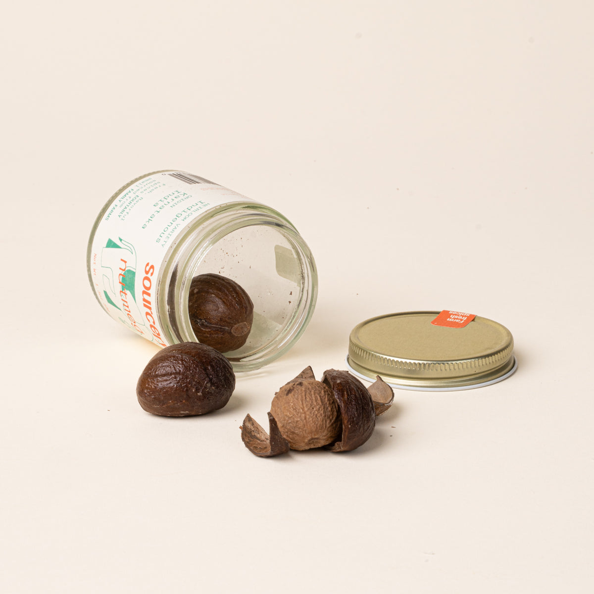 Glass jar of Sourcery nutmeg with  a cream label featuring orange lettering and a green elephant illustration, open on  its side with whole nutmeg spilling out.