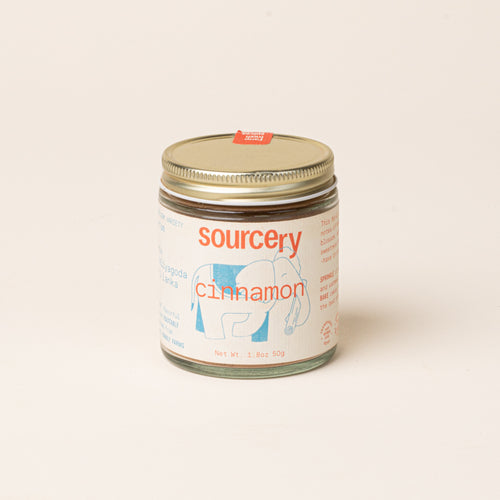 Glass jar of Sourcery cinnamon with a gold metal lid and a cream label featuring orange lettering and a blue elephant illustration.