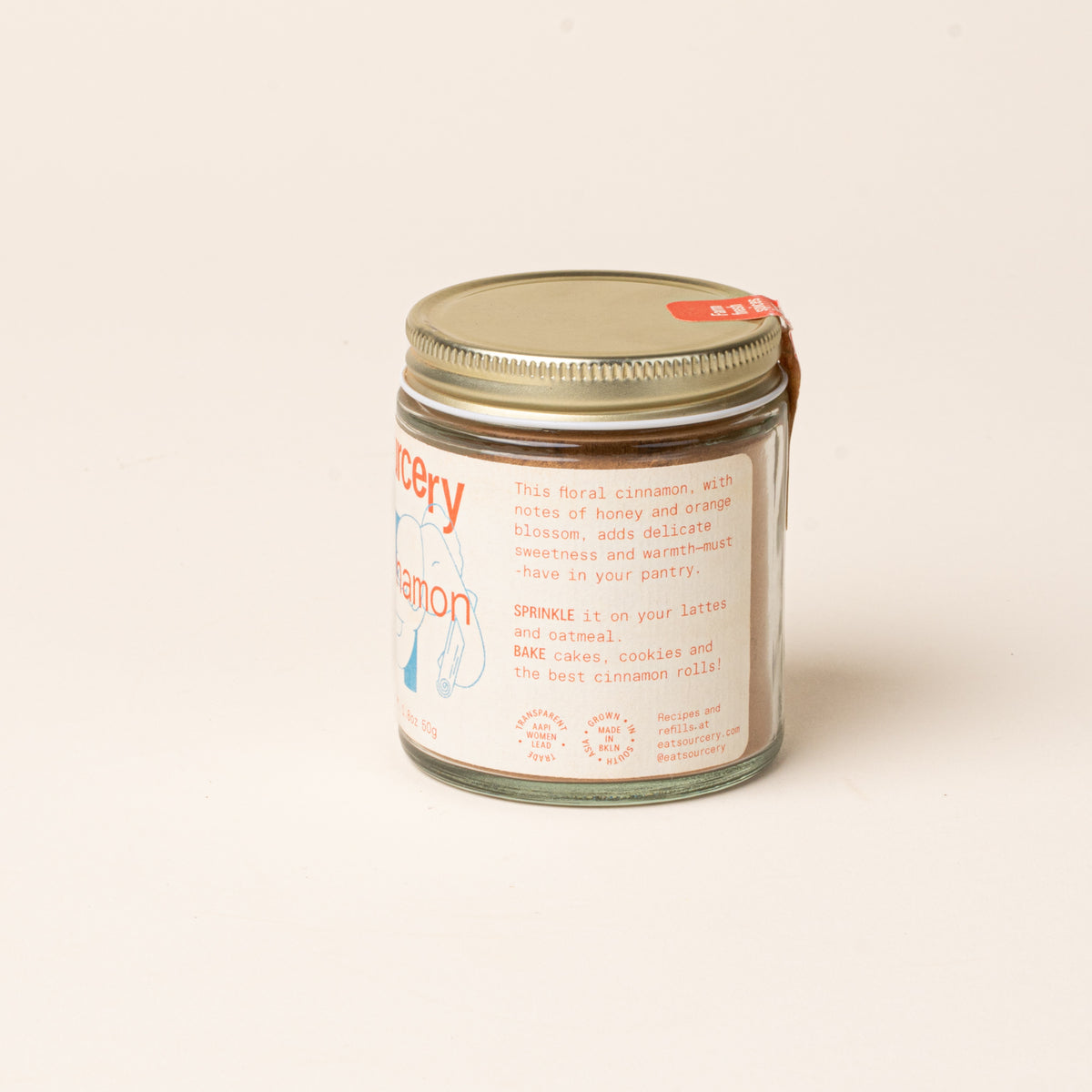 Side of Glass jar of Sourcery cinnamon with a gold metal lid and a cream label featuring orange lettering.