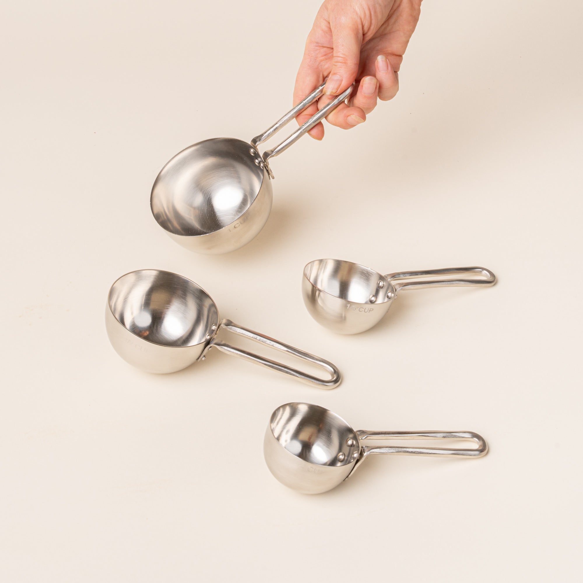 Four forged measuring cups in graduated sizes, with a hand holding one on a warm cream background.