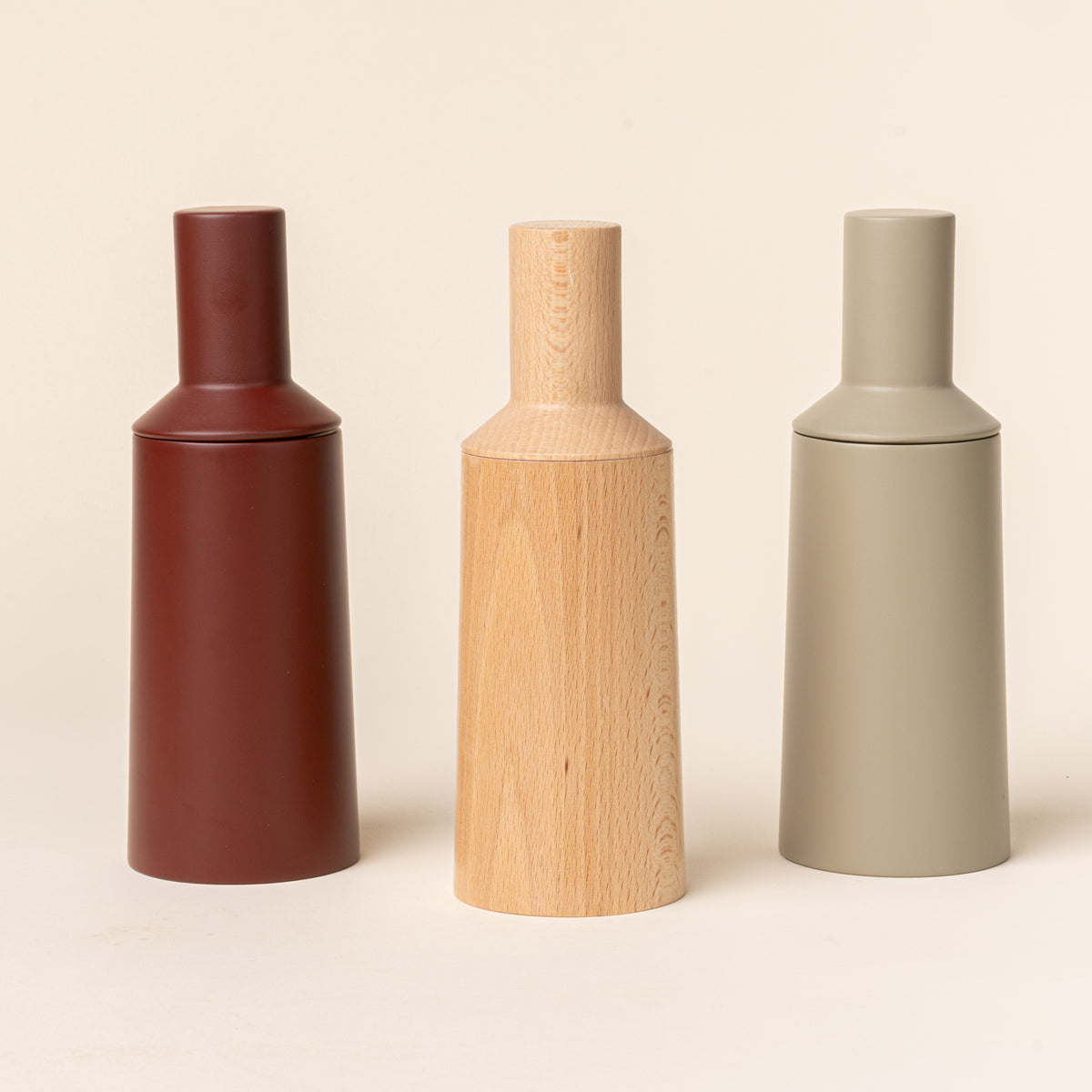 Three tall cylindrical sculptural salt and pepper grinders in dark red, natural wood, and muted light green on a light background.