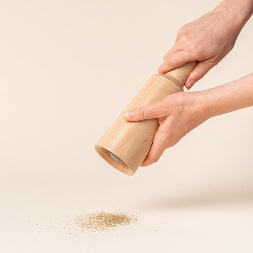 A hand holds out a tall cylindrical sculptural salt and pepper grinder in a natural light wood on a light background, milling salt