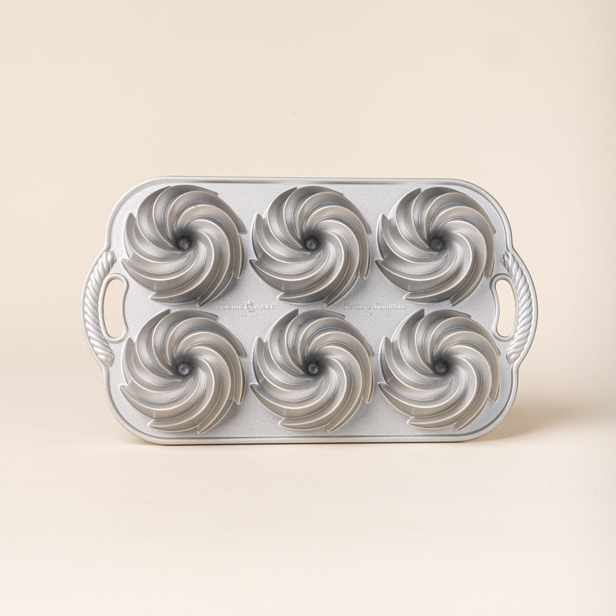 Silver metal baking pan with six spiral-shaped mini Bundt cake molds and decorative handles on each side.