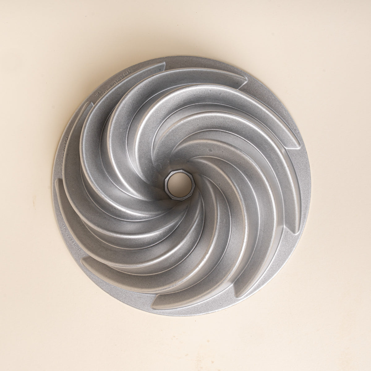 Overhead view of a silver metal Bundt cake pan with curved spiral ridges and a center opening on a cream background.