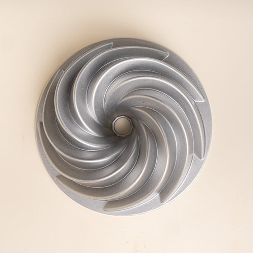 Overhead view of a silver metal Bundt cake pan with curved spiral ridges and a center opening on a cream background.