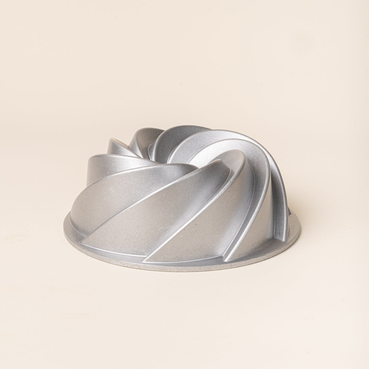 Silver metal Bundt cake pan with a sculptural spiral design and raised curved ridges on a cream background.