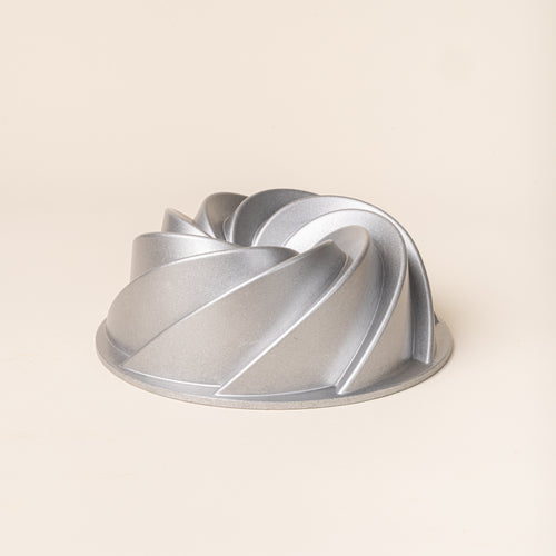 Silver metal Bundt cake pan with a sculptural spiral design and raised curved ridges on a cream background.