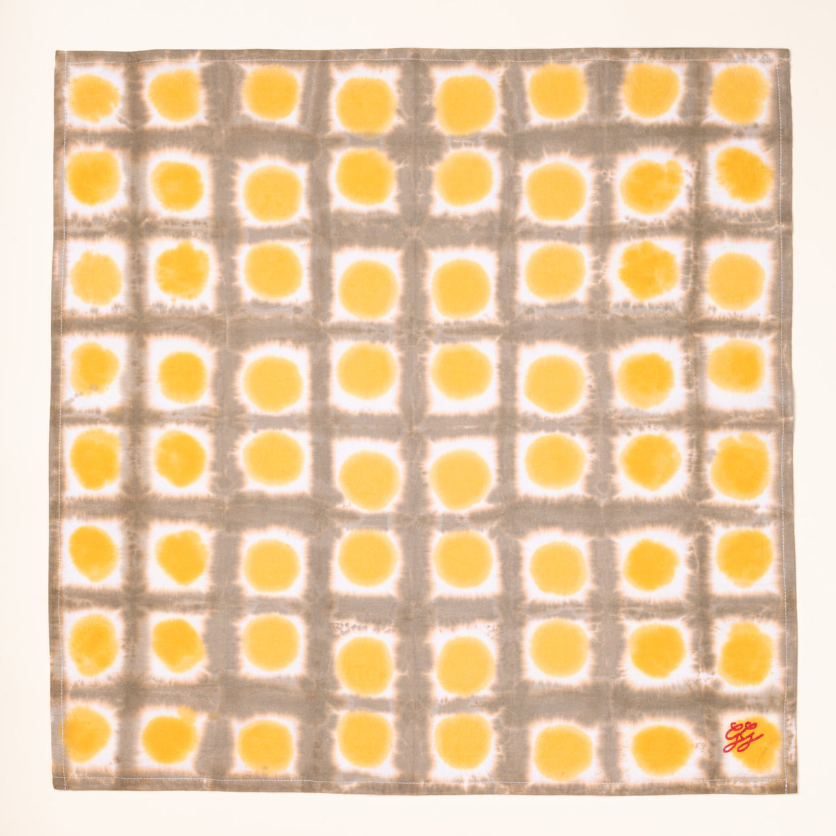 An overhead view of a flat tie-dye tea towel in a yellow colorway, with a red embroidery in the corner.