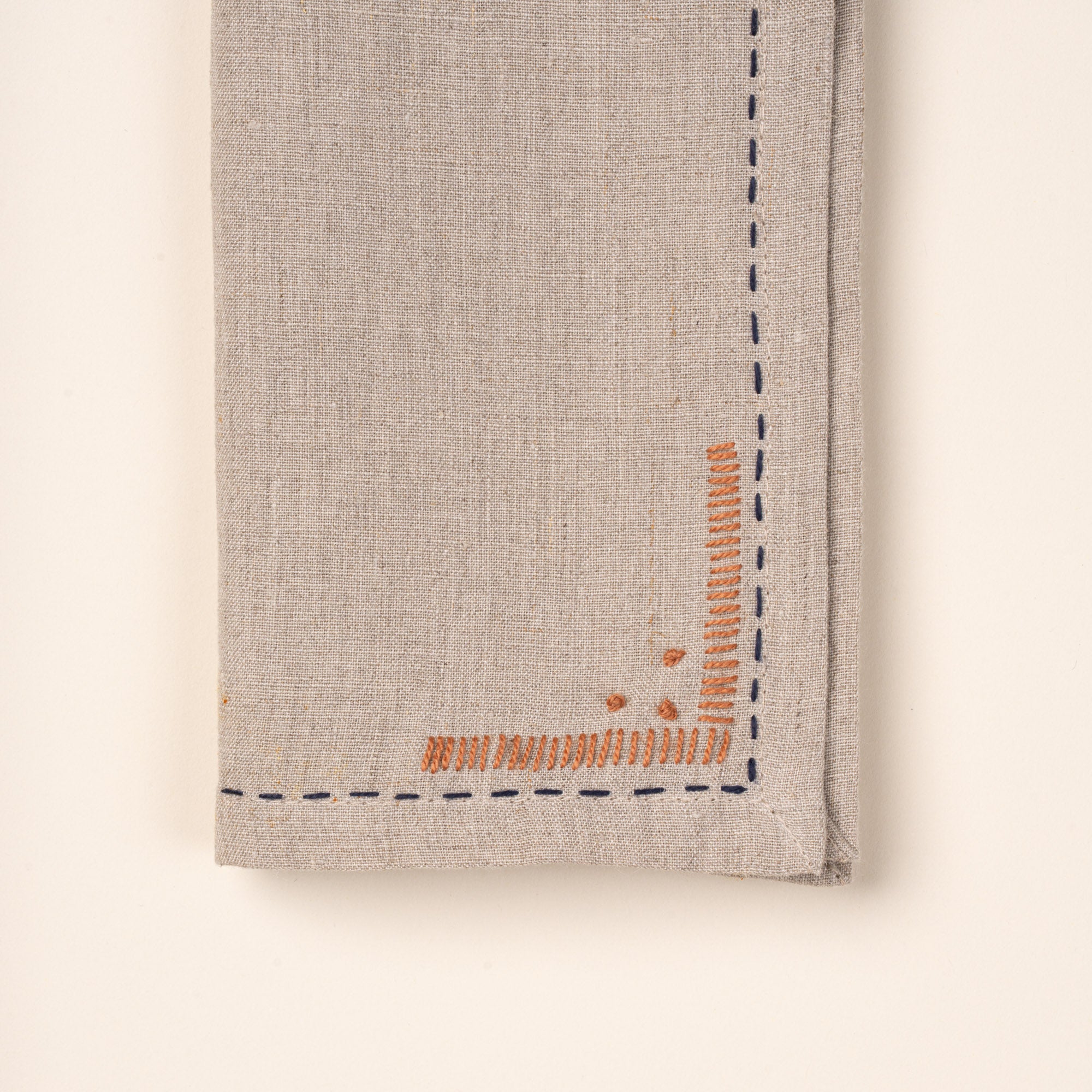 Close up of a corner of a folded natural linen napkins featuring dark blue running-stitch borders and small rust-orange embroidered corner details.