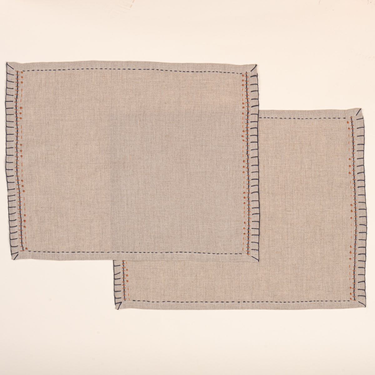 Two overlapping natural flax-colored linen placemats with hand-stitched navy borders and rust-orange accent embroidery, photographed against a warm off-white background.