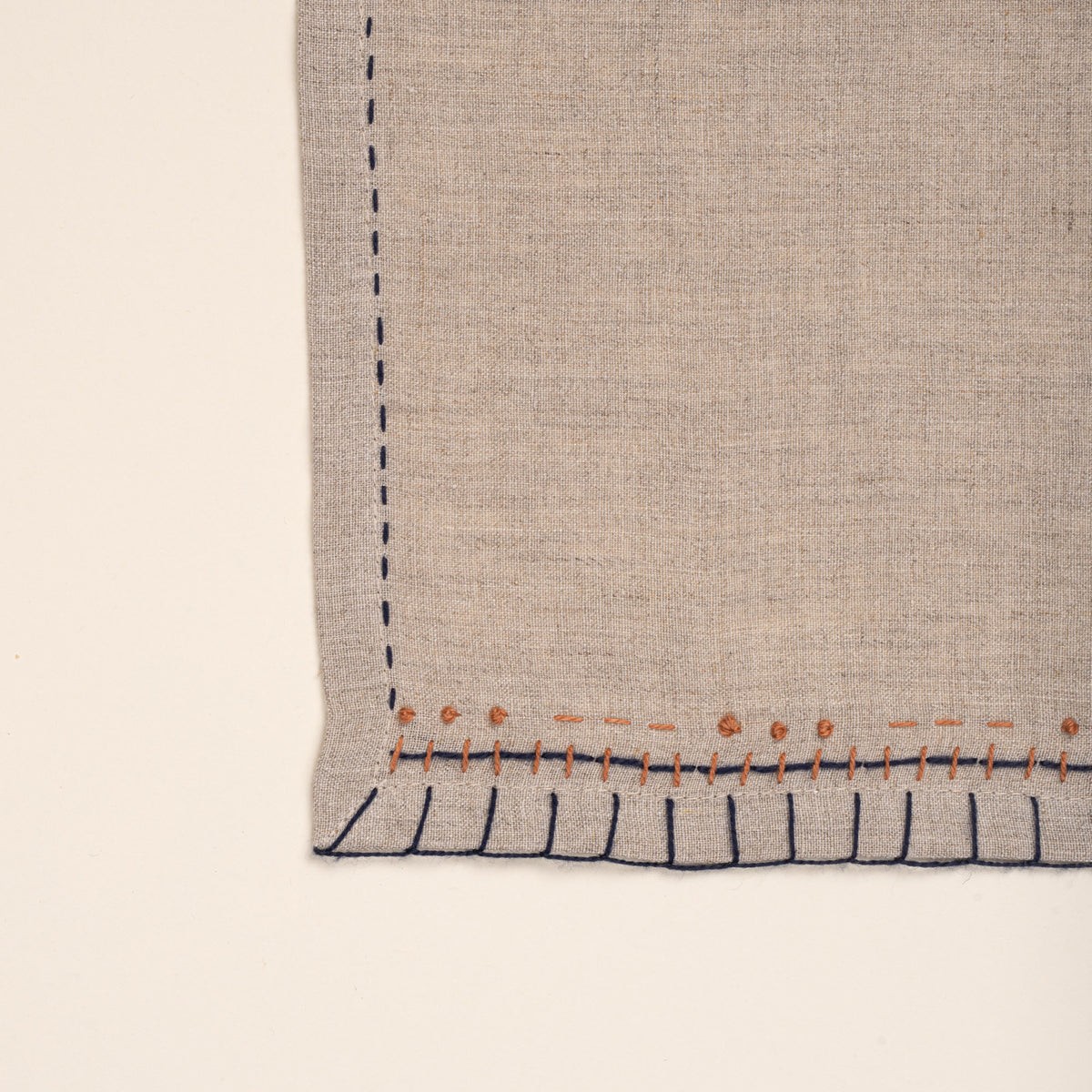 A closeup of a natural flax-colored linen placemats with hand-stitched navy borders and rust-orange accent embroidery, photographed against a warm off-white background.
