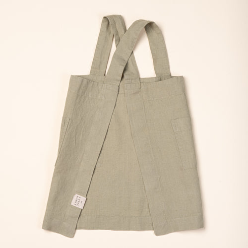 Back of a linen child aprons in light sage green with front pockets and shoulder straps.