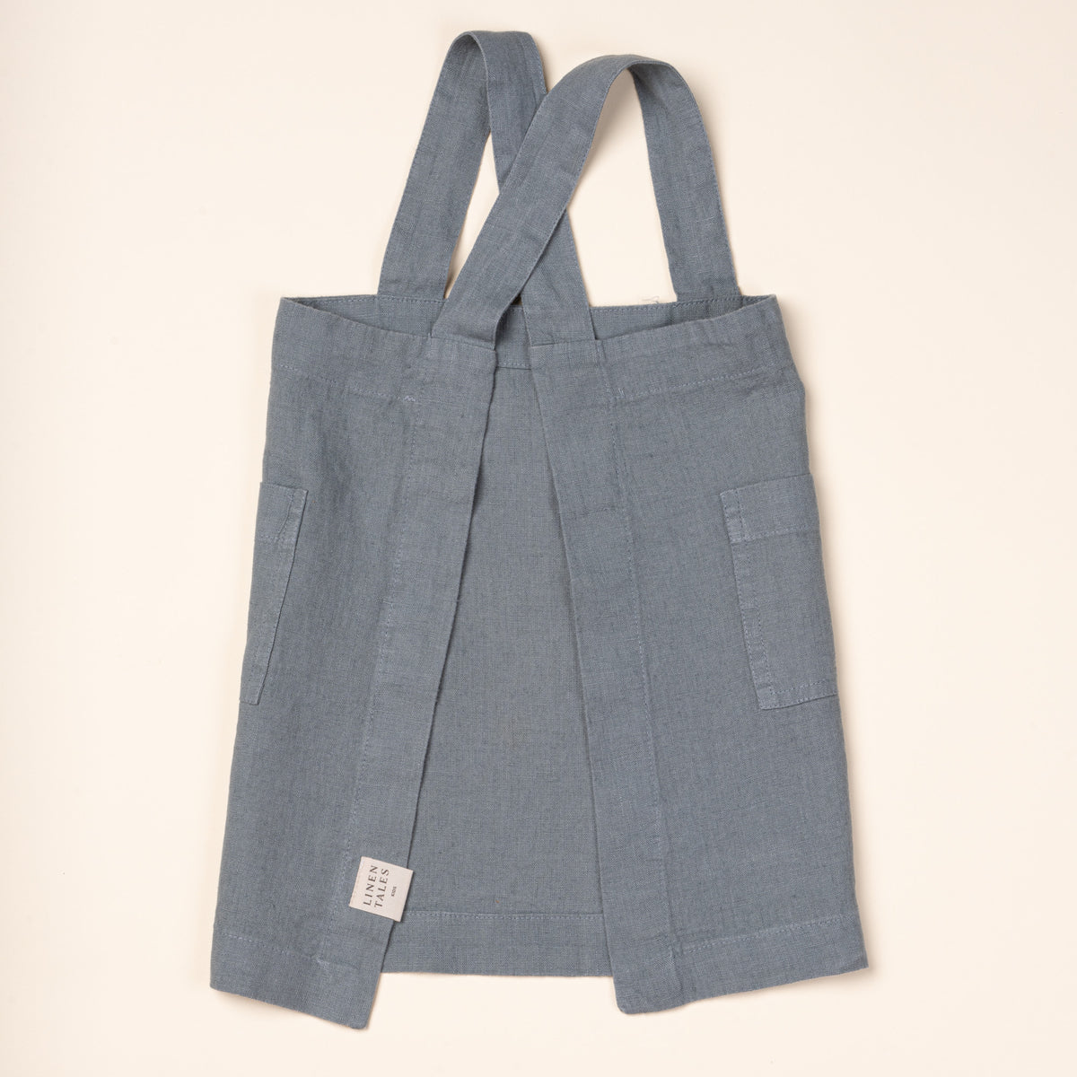 Back of a linen child apron in blue with front pockets and shoulder straps.
