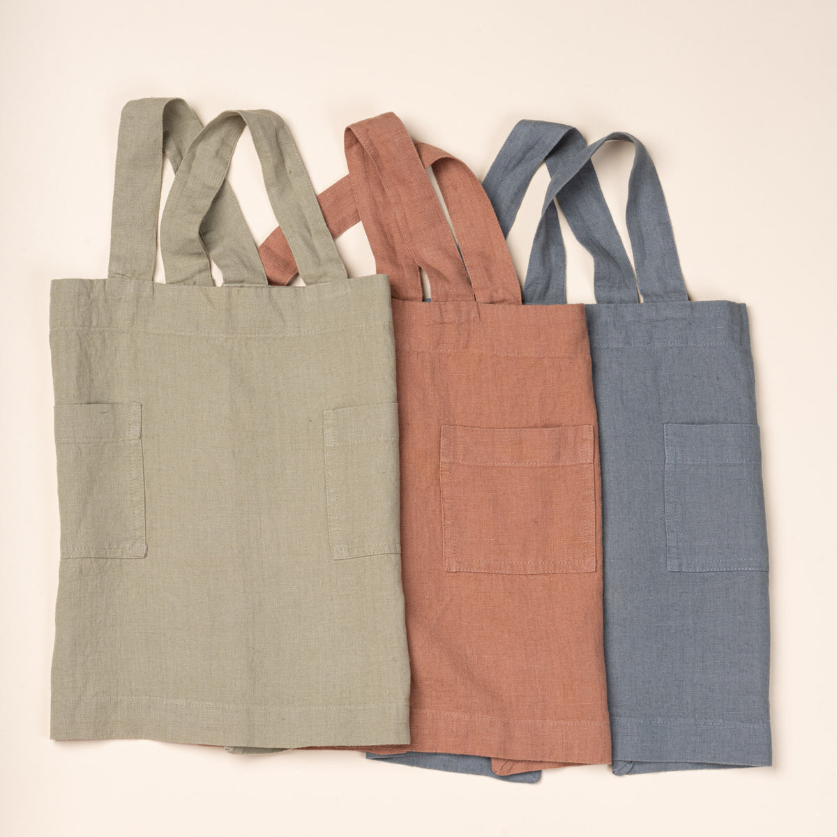 Three linen child aprons in sage green, rust, and blue with front pockets and shoulder straps.