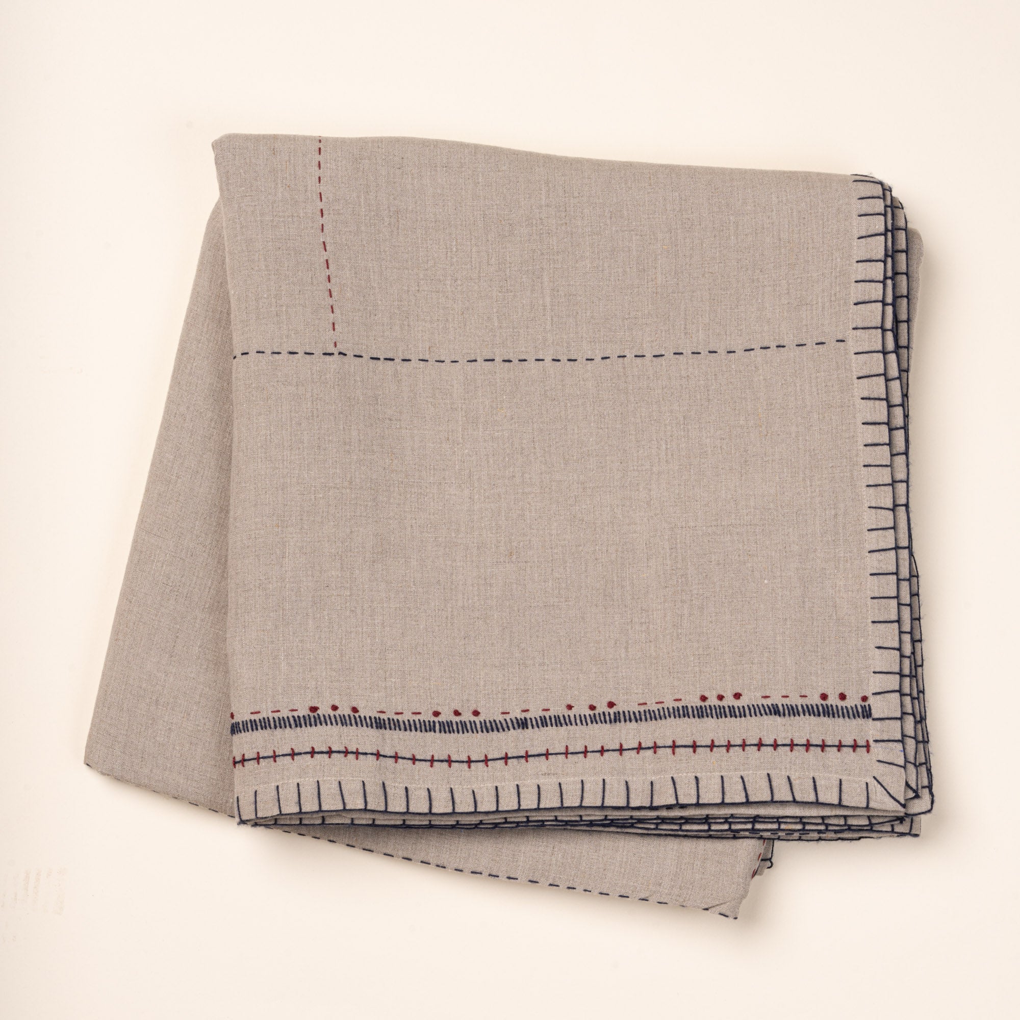 Folded natural linen tablecloth with hand-stitched navy and rust detailing and a decorative geometric border.