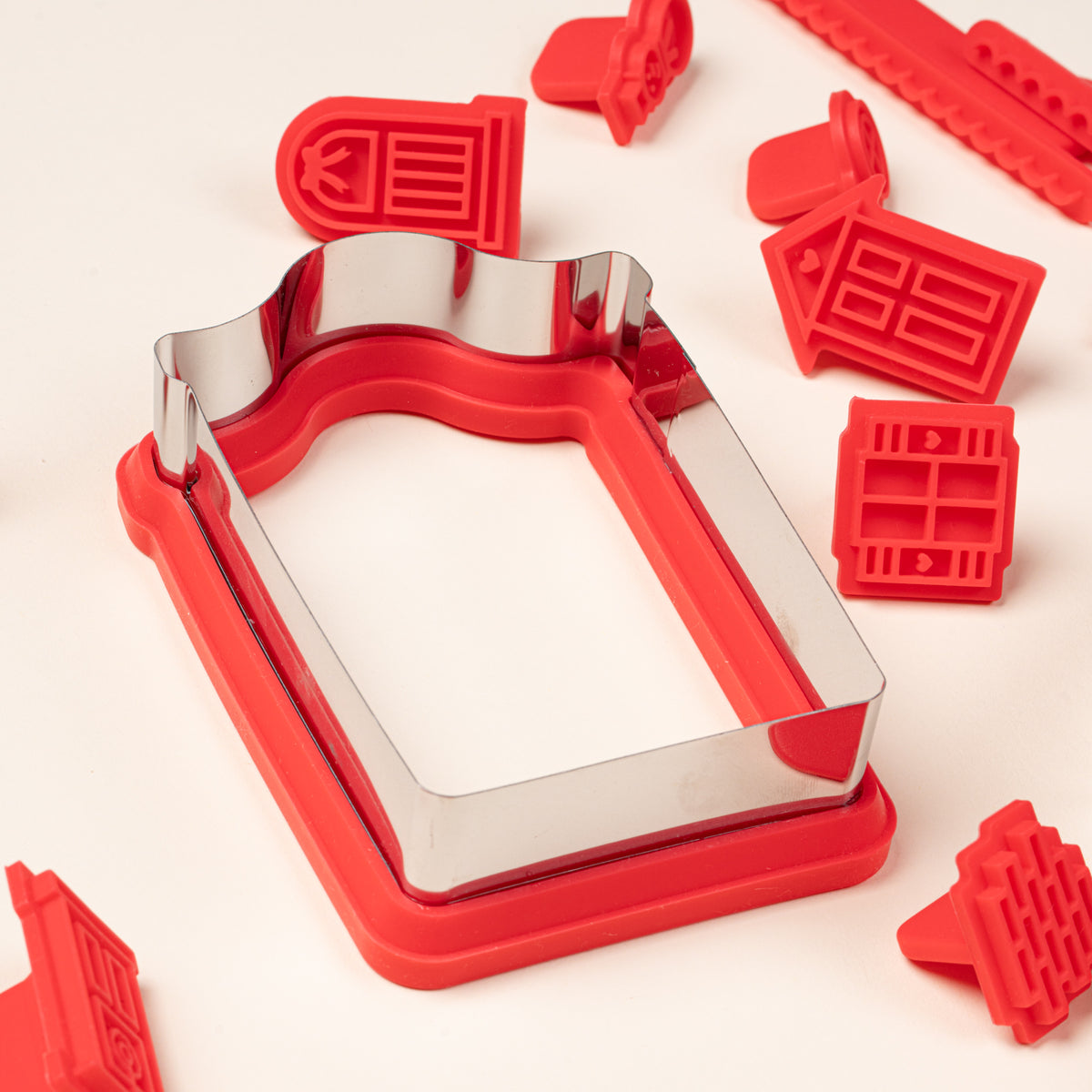 Close-up of a metal house-shaped cookie cutter with a red silicone base, surrounded by red silicone stamps featuring doors and windows.