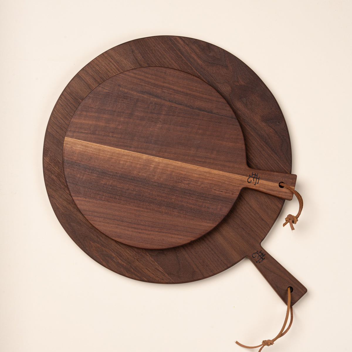 A small and large round walnut serving boards in different sizes, each with a short handle and leather hanging loop.