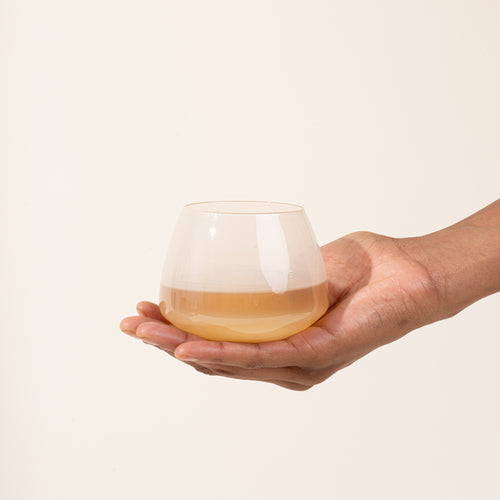 Hand holding a rounded light beige colored glass with a warm amber-colored liquid against a light neutral background.