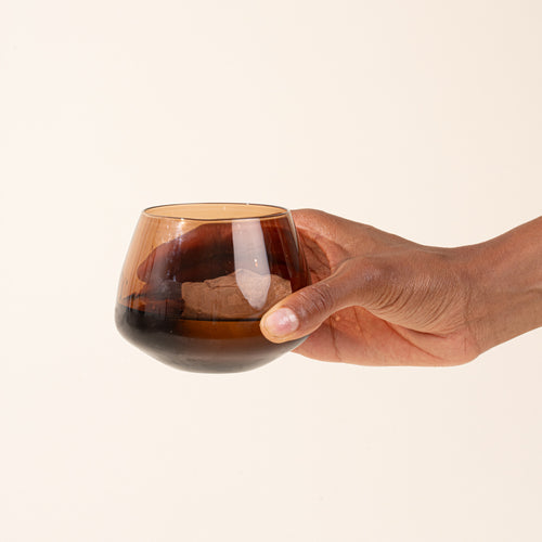 A hand holding a small, rounded stemless glass with a smoky amber-brown tint against a solid, warm off-white background.