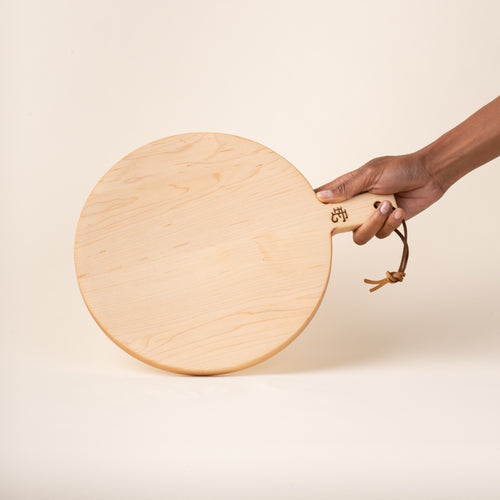 A hand holds a small round maple serving board with a short handle and leather hanging loop, shown held against a warm cream background.