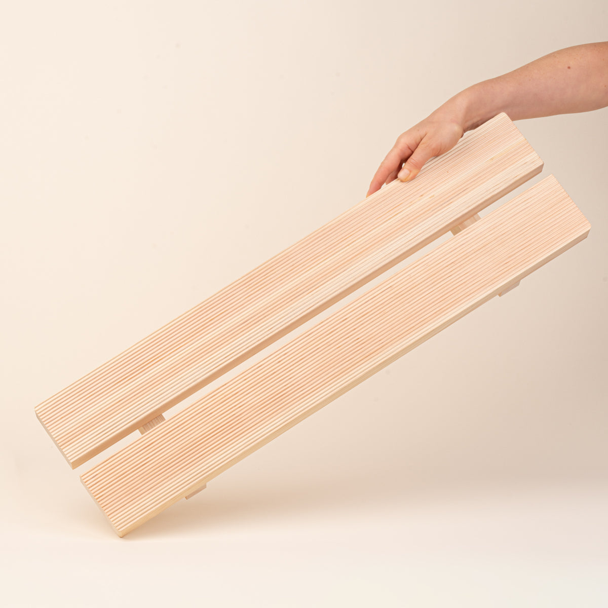 A hand holds up a long, low unfinished-wood bath tray made from two parallel planks, with simple block feet. 