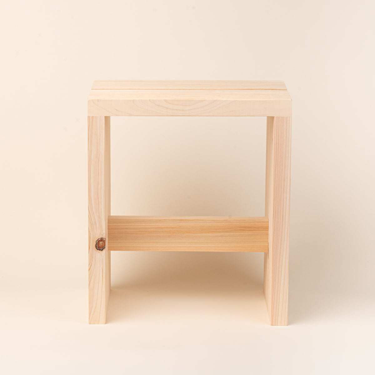 A compact unfinished-wood bath stool with thick plank construction, visible natural grain and knots, and a simple cross-brace, photographed on a warm cream background.