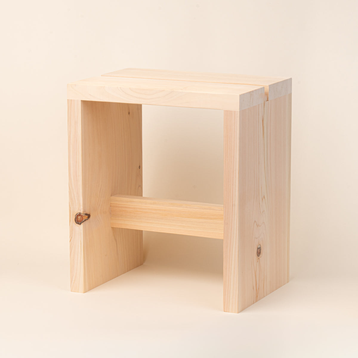 A compact unfinished-wood bath stool with thick plank construction, visible natural grain and knots, and a simple cross-brace, photographed on a warm cream background.