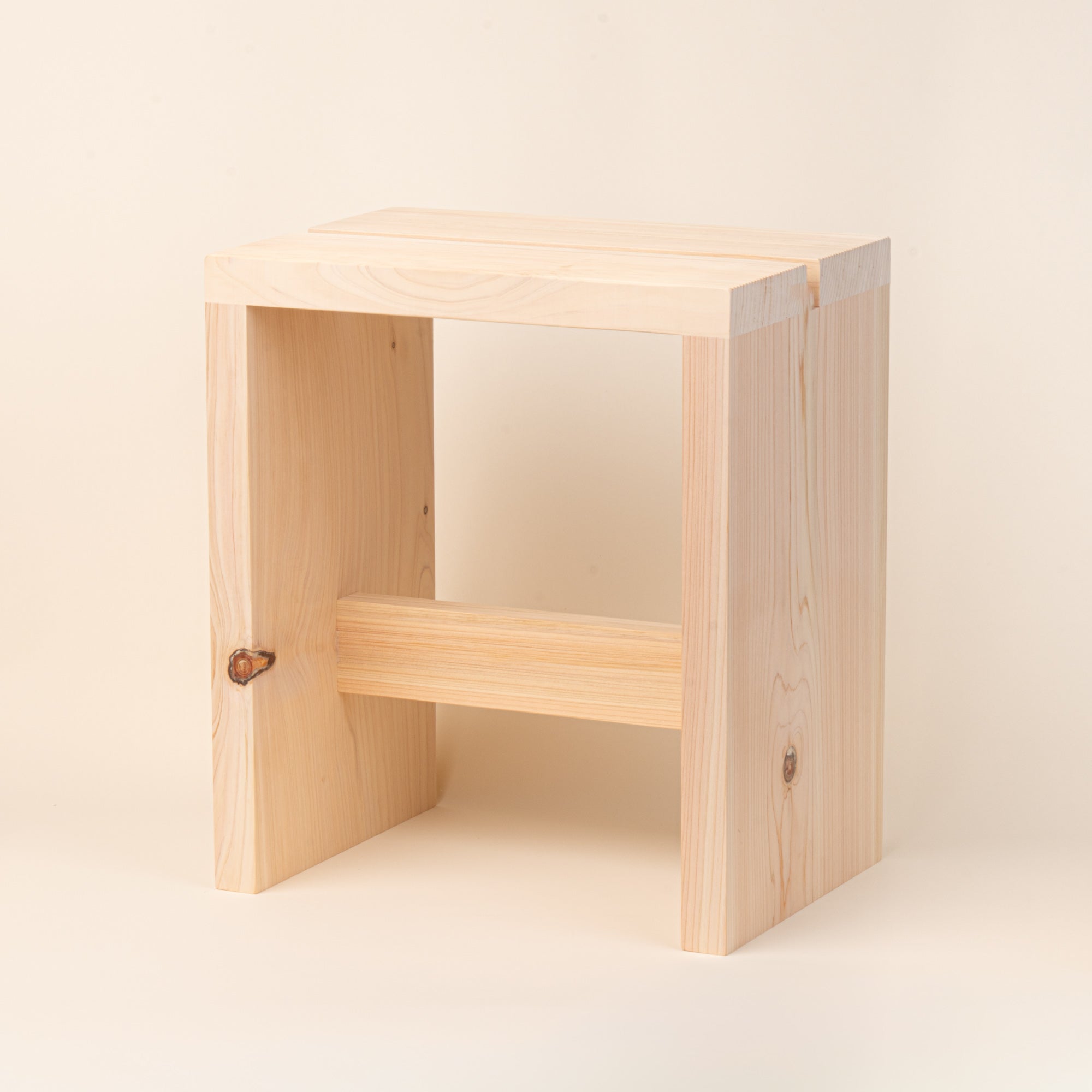 A compact unfinished-wood bath stool with thick plank construction, visible natural grain and knots, and a simple cross-brace, photographed on a warm cream background.