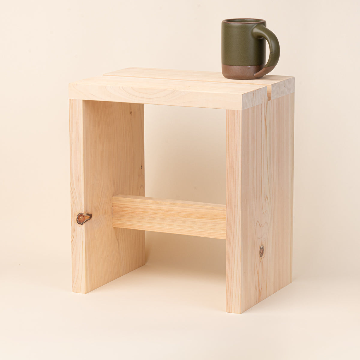 A compact unfinished-wood bath stool with thick plank construction, visible natural grain and knots, and a simple cross-brace, with an East Fork Mug on top.