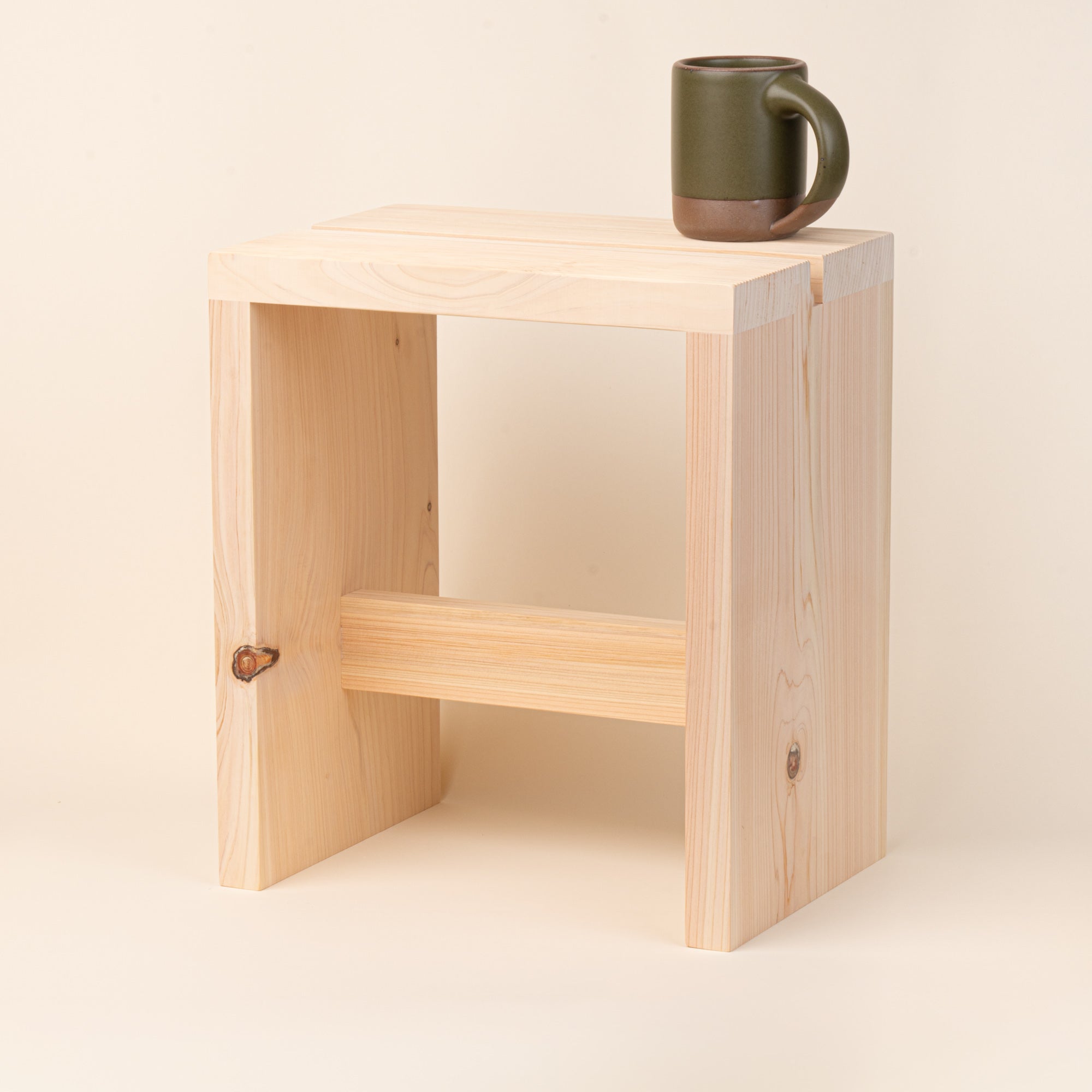 A compact unfinished-wood bath stool with thick plank construction, visible natural grain and knots, and a simple cross-brace, with an East Fork Mug on top.