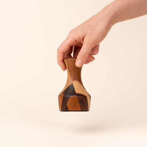 A hand holds a sculptural wooden biscuit cutter with a tapered neck, flared top, and geometric patchwork of light and dark wood tones.