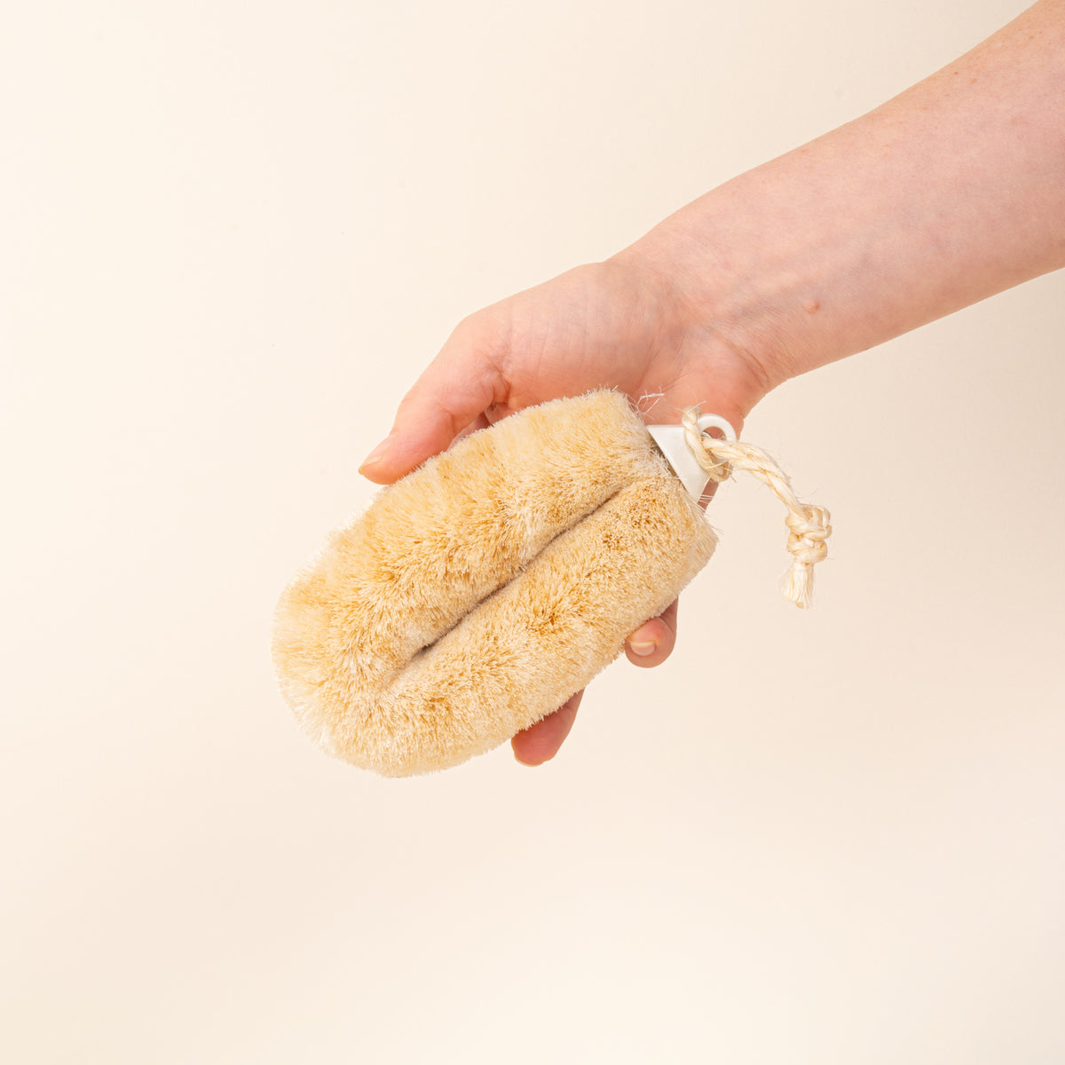 A hand holds a natural-fiber body or bath brush with dense cream-colored bristles and a braided hanging loop, photographed on a warm cream background.