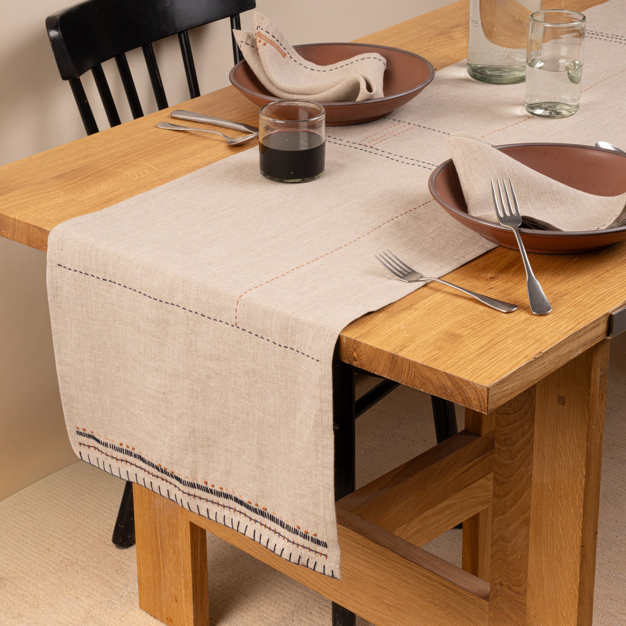 A natural linen table runner styled across a warm oak dining table with matching napkins, dark ceramic dinnerware, simple glassware, and silver flatware.