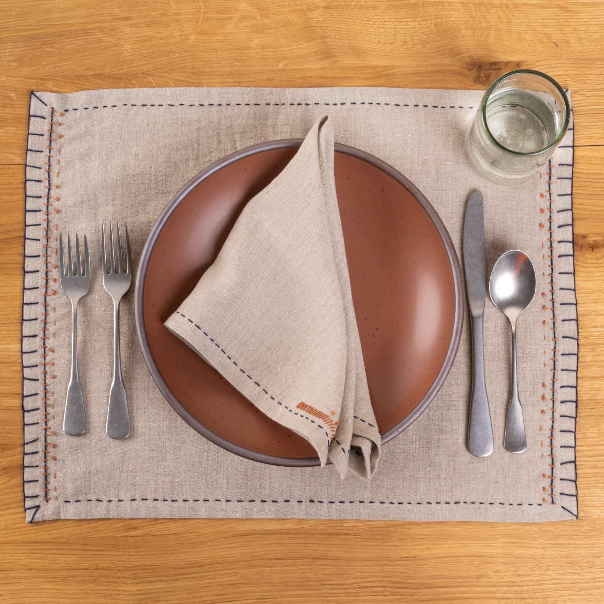 A natural linen placemat and coordinating napkin with dark blue running-stitch borders and rust-orange embroidered details, styled with a warm terracotta ceramic plate, silver flatware, and a clear glass on a wood tabletop.