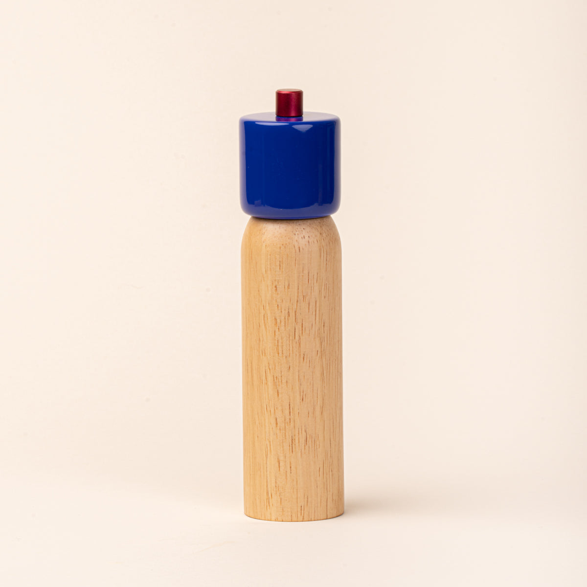 Wooden pepper mill with a glossy cobalt-blue top and red knob, photographed against a warm cream background.