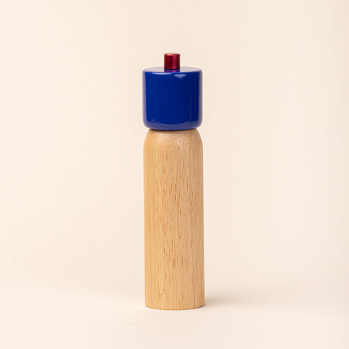 Wooden pepper mill with a glossy cobalt-blue top and red knob, photographed against a warm cream background.