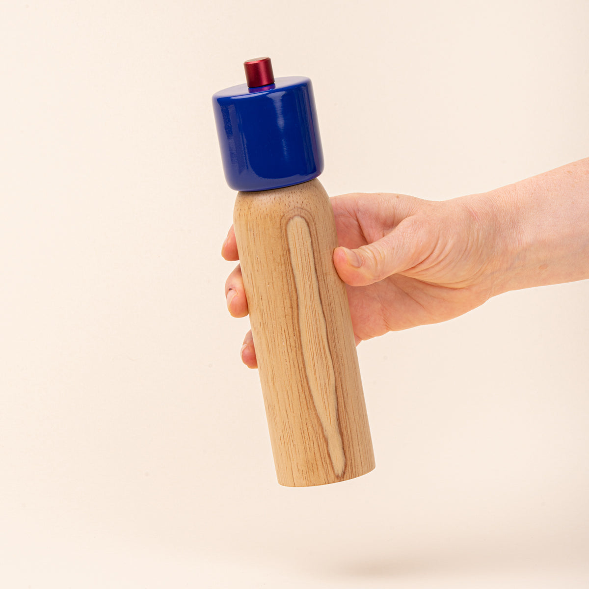 A hand holds a wooden pepper mill with a glossy cobalt-blue top and red knob, photographed against a warm cream background.