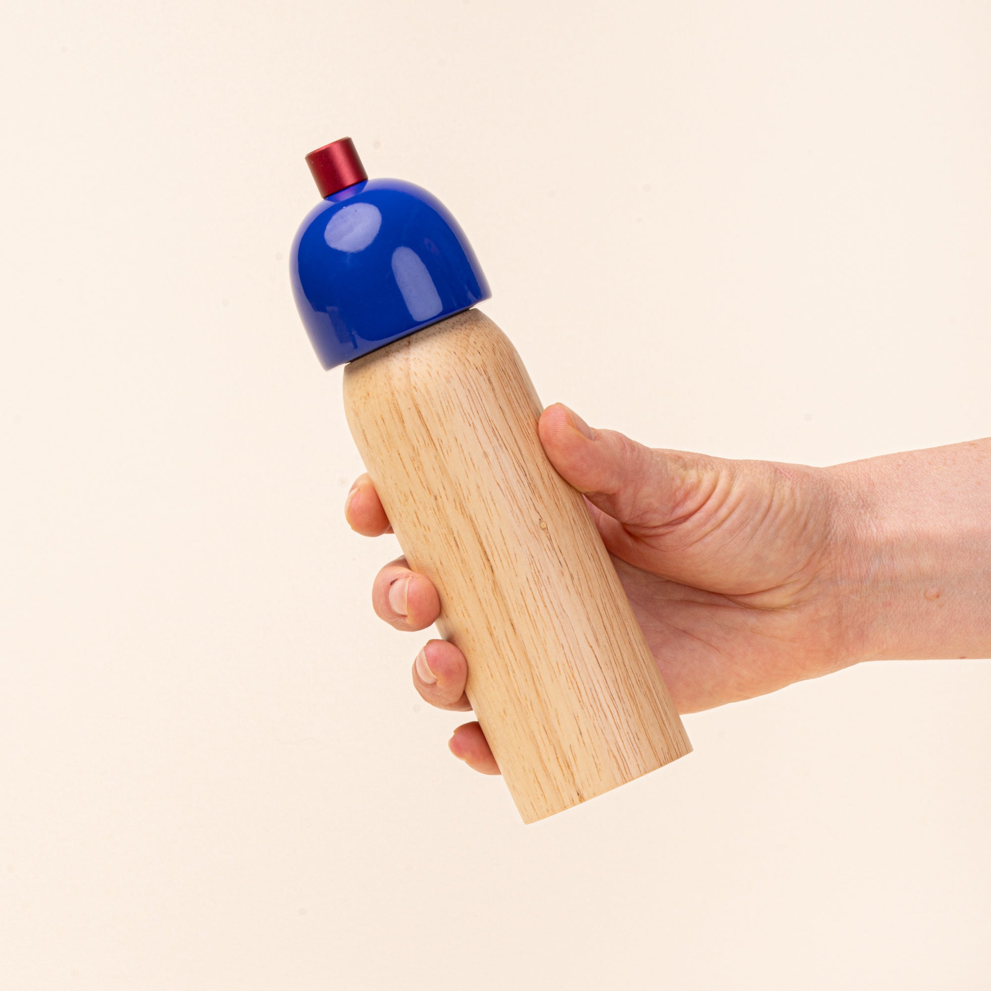 A hand holds a wooden salt mill with a glossy cobalt-blue top and red knob, photographed against a warm cream background.