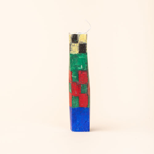 A tall, tapered rectangular candle decorated with a hand-painted geometric pattern in cobalt blue, red, green, yellow, and black, with a white wick at the top.