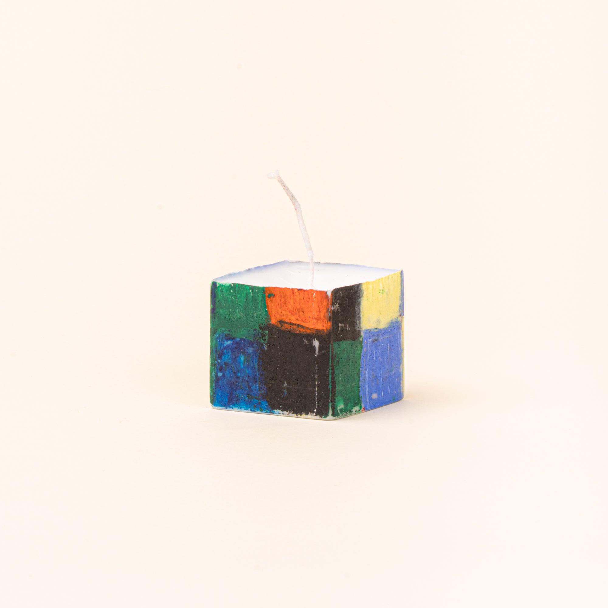 A tall, hand-painted rectangular candle shown beside its original green-and-white checkerboard paper packaging. The candle features blocks of cobalt blue, red, green, yellow, and black, with a white wick at the top.