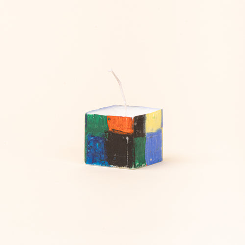 A tall, hand-painted rectangular candle shown beside its original green-and-white checkerboard paper packaging. The candle features blocks of cobalt blue, red, green, yellow, and black, with a white wick at the top.